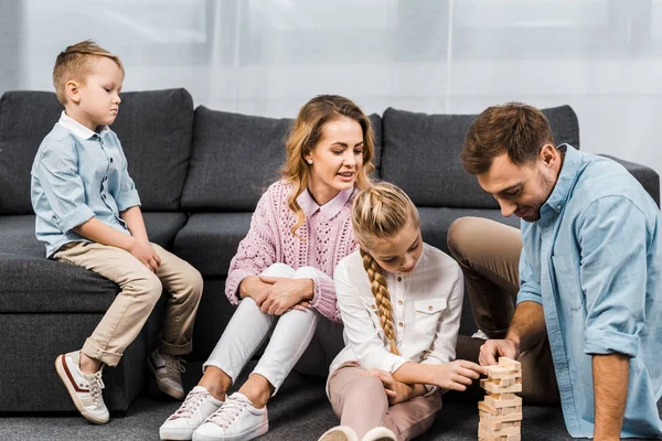Двое счастливых родителей играют в блоки с дочкой на полу, а расстроенный мальчик сидит на диване в гостиной — стоковое фото
