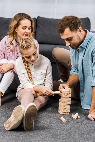 Dois pais felizes jogando blocos de madeira torre jogo com a filha no chão na sala de estar — Fotografia de Stock