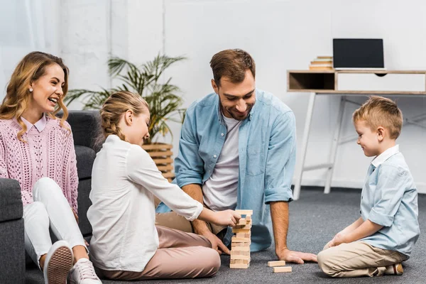 Two happy parents playing blocks wood tower game with daughter and son on floor in living room — Stock Photo