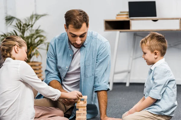 Padre jugando bloques de madera juego de torre con hija e hijo en el suelo en la sala de estar - foto de stock