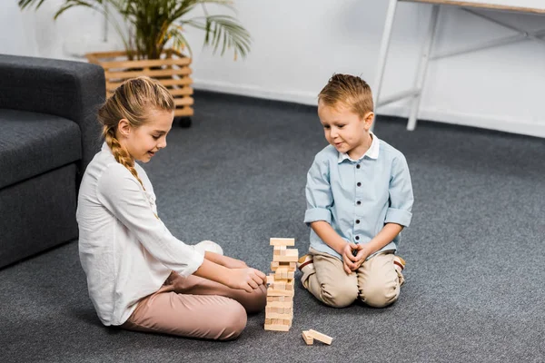 Lindos hermanos sentados en el suelo y jugando bloques torre de madera juego en la sala de estar - foto de stock