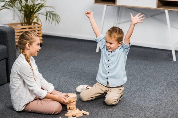 Niño feliz con las manos levantadas y linda chica sentada en el suelo y jugando bloques de madera juego de torre en el apartamento - foto de stock