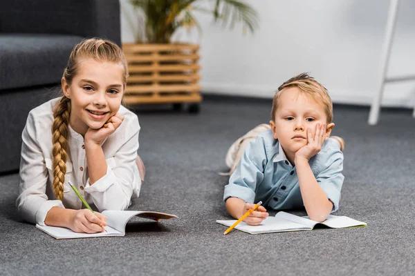 Niedliche Mädchen und Jungen liegen auf dem Boden, schreiben in Notizbücher und schauen in die Kamera im Wohnzimmer — Stockfoto