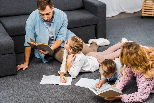 Батьки читають книги, коли діти пишуть в блокнотах на підлозі в квартирі — стокове фото