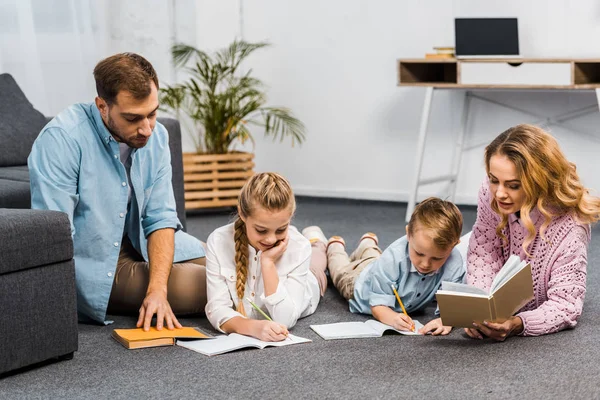 Parents reading books while siblings writing in exercise books on floor in apartment — Stock Photo