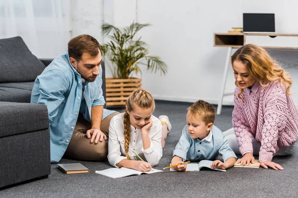 Parents looking at girl and boy lying on floor and writing in exercise books in apartment — Stock Photo