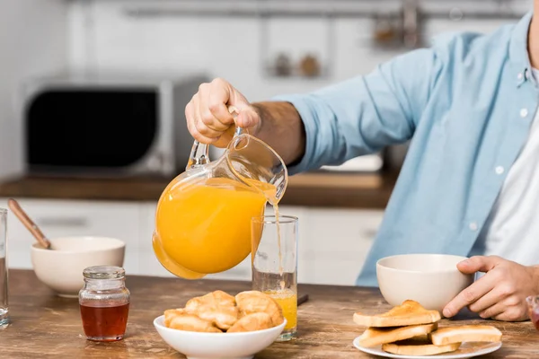 Cropped view of man pouring orange juice in glass on kitchen table — Stock Photo