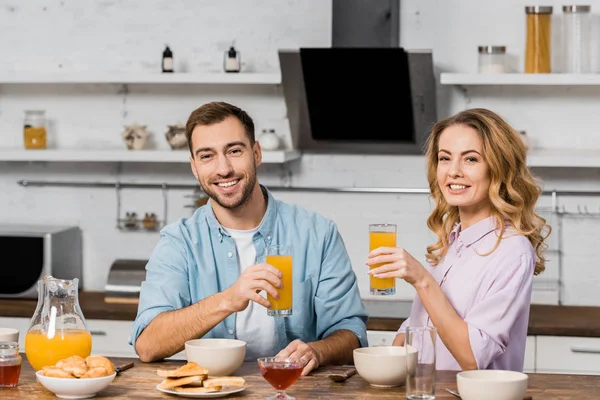 Smiling woman and man sitting at kitchen table and holding glasses with orange juice — Stock Photo