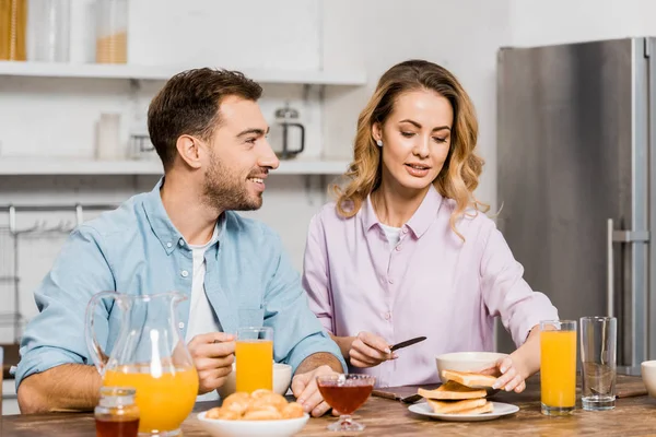Hombre guapo mirando mujer bonita sosteniendo cuchillo y tostadas en la cocina - foto de stock