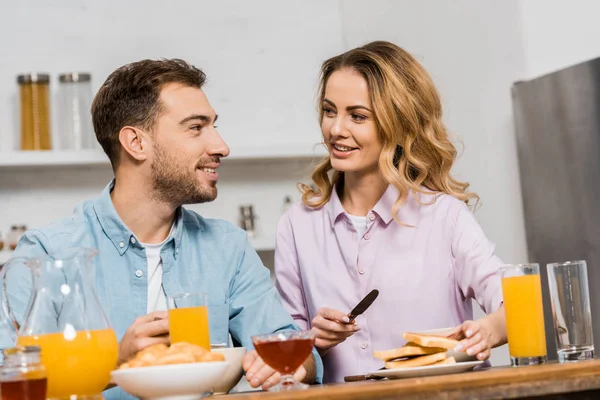 Mujer bonita sosteniendo cuchillo y tostadas y mirando marido guapo en la mesa - foto de stock