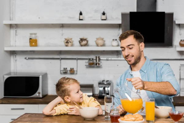 Menino bonito olhando para o pai sorrindo derramando suco de laranja em vidro na mesa da cozinha — Fotografia de Stock