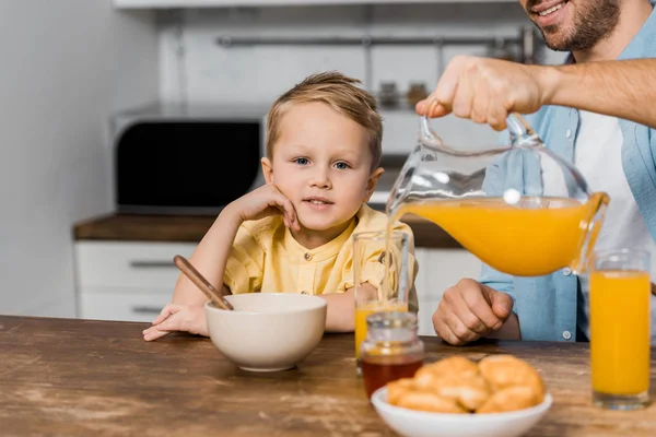 Recortado vista de sonriente padre verter jugo de naranja en vidrio y lindo chico mirando a la cámara - foto de stock