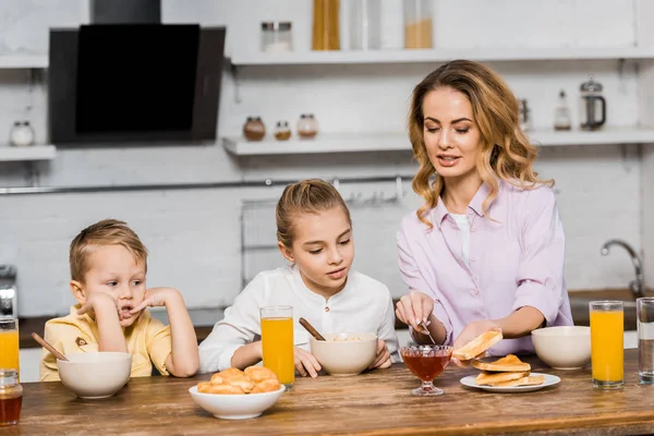 Cute siblings looking at pretty woman spreading jam on toast at table — Stock Photo