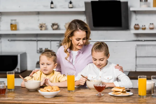 Hübsche Frau umarmt niedliche Kinder, die Haferflocken am Küchentisch essen — Stockfoto