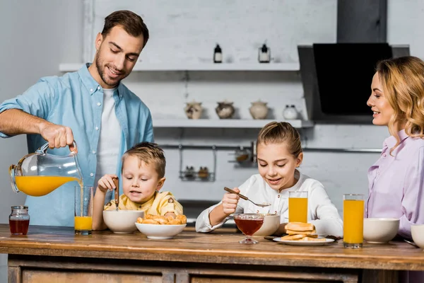 Bello padre versando succo d'arancia in vetro mentre i bambini mangiano farina d'avena a tavola — Foto stock