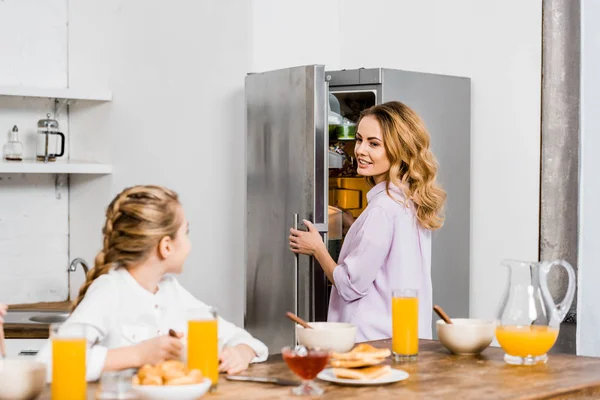 Menina sentada à mesa e olhando para a mãe abertura frigorífico — Fotografia de Stock