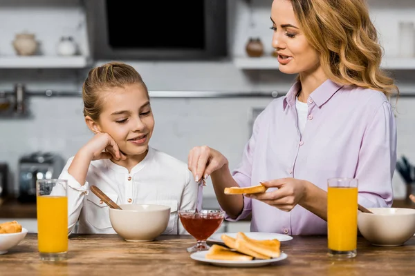 Sorridente ragazza guardando bella donna diffusione marmellata sul pane tostato in cucina — Foto stock