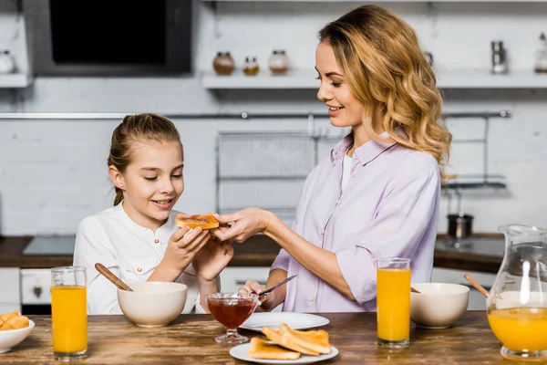 Sonriente mujer dando tostadas con mermelada a linda hija en la mesa en la cocina - foto de stock