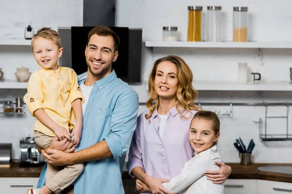 Guapo hombre sosteniendo hijo y bonita madre abrazando linda hija en la cocina - foto de stock