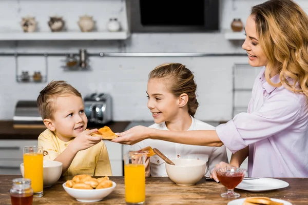 Sonriente chica mirando pequeño hermano tomando tostadas con mermelada de madre en cocina - foto de stock
