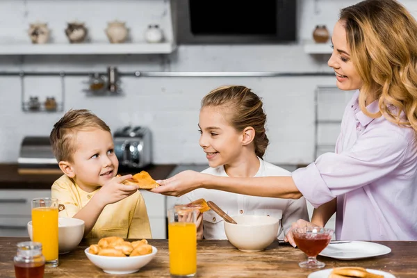 Fille souriante regardant petit frère portant un toast avec de la confiture de la mère dans la cuisine — Photo de stock