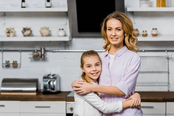 Atractiva mujer y linda hija abrazando y mirando a la cámara en la cocina - foto de stock