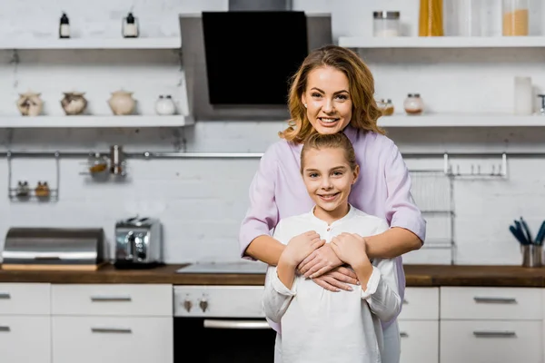Улыбающаяся женщина обнимает симпатичную дочь и смотрит в камеру на кухне — стоковое фото
