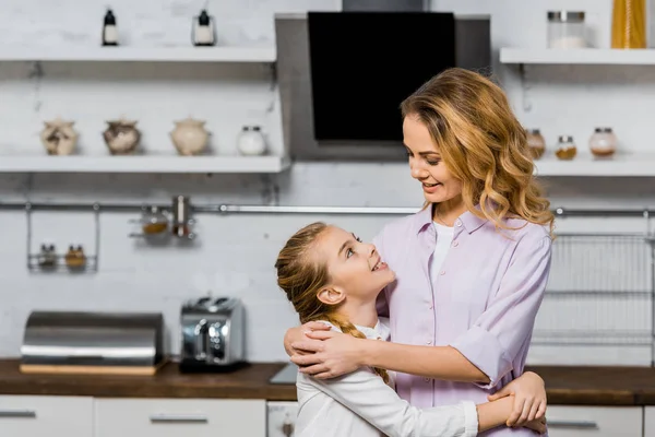 Bonita mujer y linda hija abrazándose y mirándose en la cocina - foto de stock
