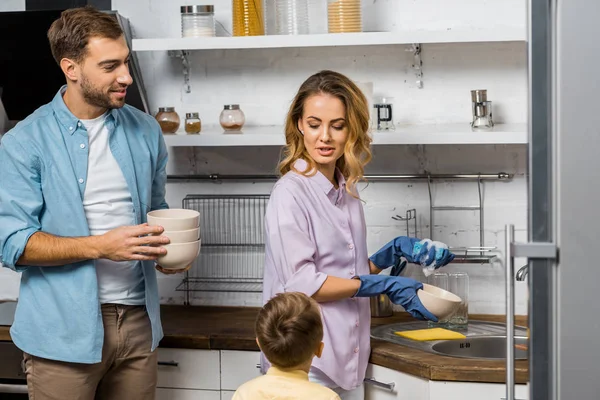 Hombre guapo sosteniendo cuencos y mirando bonita esposa lavando platos y hablando con hijo - foto de stock