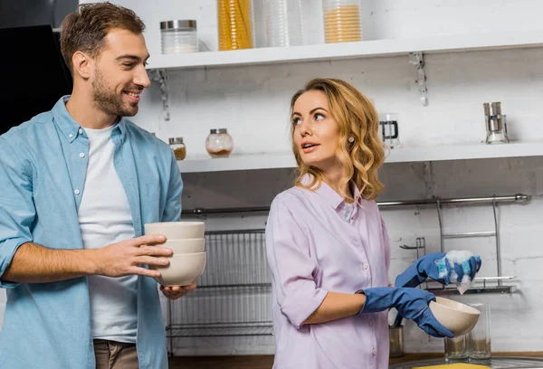 Attraktive Frau in Gummihandschuhen, die Geschirr spült und lächelnden Mann mit Schalen in der Küche ansieht — Stockfoto