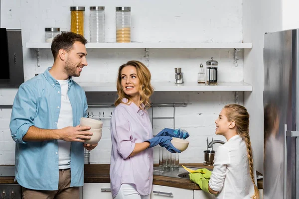 Bonita mujer e hija en guantes de goma lavando y mirando guapo padre sosteniendo cuencos en la cocina - foto de stock