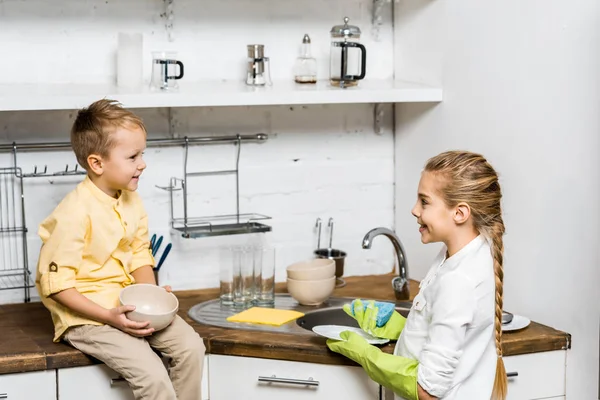 Cute girl in rubber gloves washing dishes and looking at smiling boy sitting on table and holding bowl in kitchen — Stock Photo