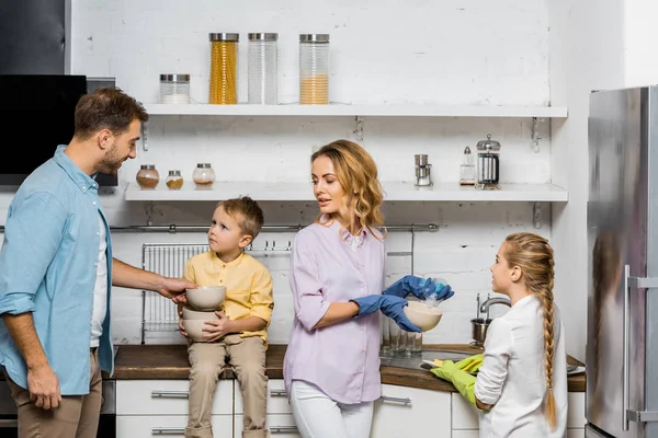 Madre e hija en guantes de goma lavándose mientras padre e hijo sostienen tazones en la cocina - foto de stock