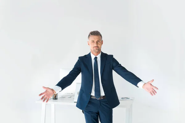 Красивый бизнесмен, стоящий с распростертыми объятиями возле стола с компьютером, изолированным на белом — стоковое фото