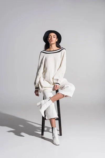 Attractive african american girl in stylish white clothes and hat sitting on chair and looking at camera on white — Stock Photo