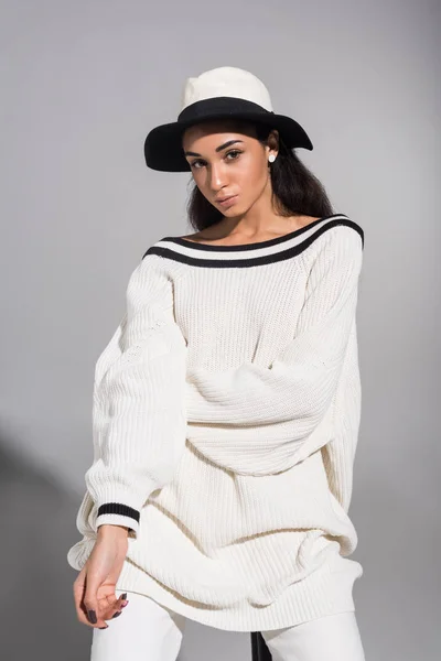 Attraente modello afroamericano femminile in eleganti vestiti bianchi e cappello seduto sulla sedia e guardando la fotocamera su bianco — Foto stock