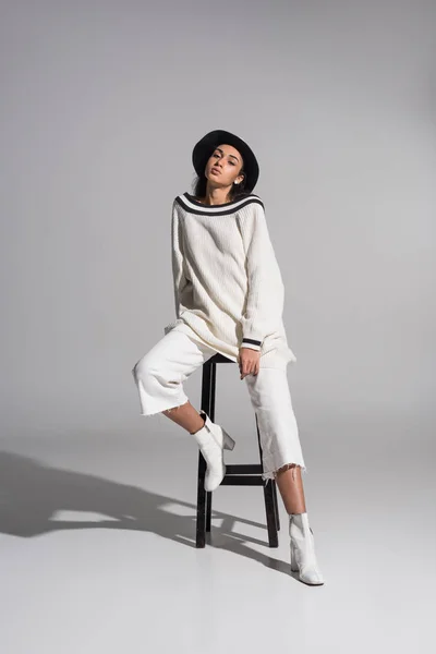 Full length of attractive african american girl in stylish white clothes and hat sitting on chair and looking at camera on white — Stock Photo