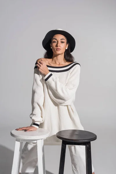 Attractive african american girl in stylish white clothes and hat standing near black and white chairs and looking at camera on white — Stock Photo