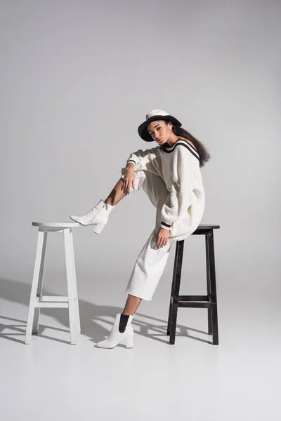 Attractive african american girl in fashionable clothes and hat sitting on black and white chairs on white — Stock Photo