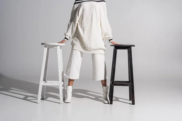 Cropped image of girl in fashionable sweater and trousers standing between black and white chairs on white — Stock Photo