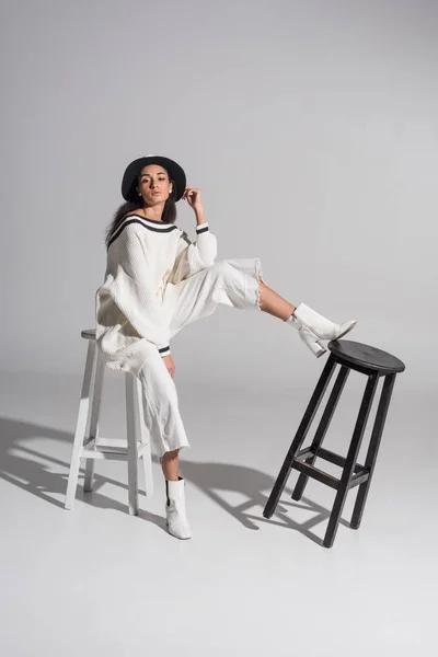 Attrayant afro-américain fille en vêtements blancs élégants et chapeau assis sur chaise blanche et regardant la caméra sur blanc — Photo de stock