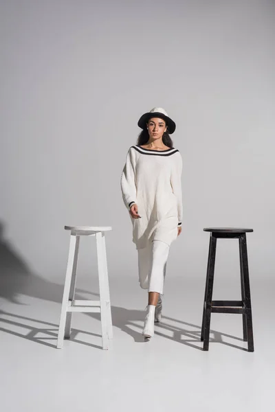 Jolie fille afro-américaine en vêtements blancs élégants et chapeau marchant entre les chaises noires et blanches sur blanc — Photo de stock