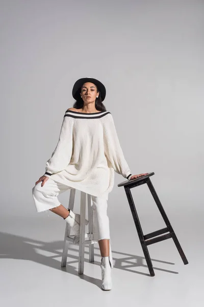 Attraente ragazza afroamericana in eleganti vestiti bianchi e cappello in posa su sedie in bianco e nero su bianco — Foto stock