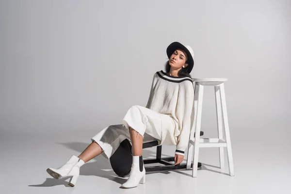 Attractive african american girl in stylish white clothes and hat posing with black and white chairs and looking at camera on white — Stock Photo