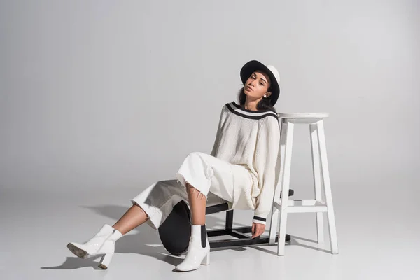 Attrayant afro-américain fille en vêtements blancs élégants et chapeau posant avec des chaises noires et blanches et en regardant la caméra sur blanc — Photo de stock