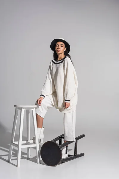 Jolie fille afro-américaine dans des vêtements blancs élégants et chapeau debout sur des chaises noires et blanches sur blanc — Photo de stock