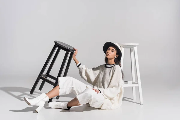 Attractive african american girl in stylish white clothes and hat sitting near black and white chairs on white — Stock Photo