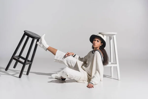 Belle fille afro-américaine en vêtements blancs à la mode et chapeau assis près de chaises noires et blanches sur blanc — Photo de stock