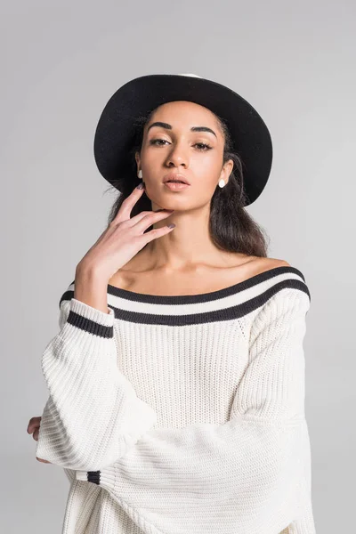 Attractive african american girl in stylish white sweater and hat touching face and looking at camera isolated on white — Stock Photo