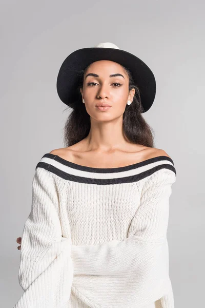 Приваблива афроамериканська дівчина в стильному білому светрі і капелюсі дивиться на камеру ізольовано на білому — стокове фото